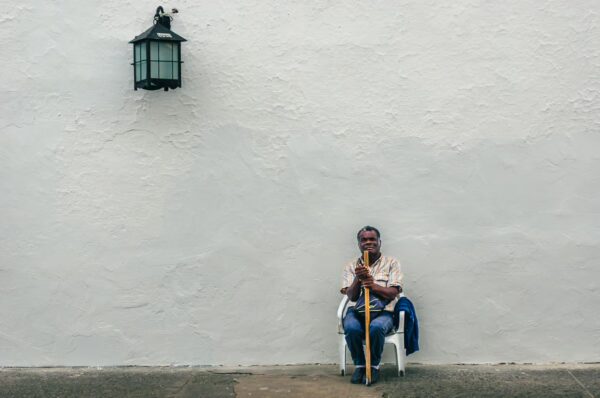 A man sitting on a chair against a white wall in Popayán, Cauca, Colombia, holding a staff next to a lantern.