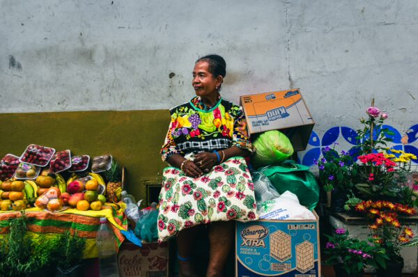Vibrant fruits and flowers are displayed at a market in San Antonio, Valle del Cauca, Colombia, with a woman wearing a colorful dress.