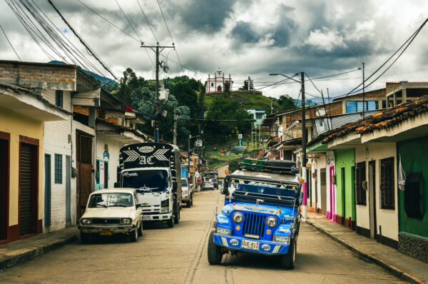 Vibrant street in Cauca, Colombia, lined with colorful houses, parked vehicles, and a distant hillside with a church.