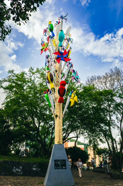 Colorful mobile sculpture with vibrant birds and pinwheels amidst lush greenery at Colina de San Antonio, Cali, Valle del Cauca.