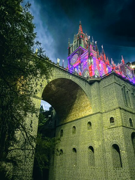 Las Lajas Sanctuary towers dramatically over the arching stone bridge, illuminated in vibrant colors against a dark sky.