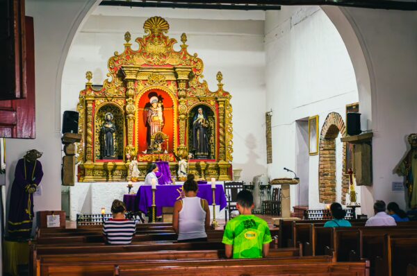 Altar with intricate golden details and religious statues, vibrant purple cloth, and parishioners seated in a church in Cali, Valle del Cauca.