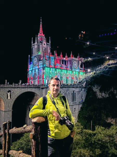 Las Lajas Sanctuary illuminated at night, showcasing vibrant colors with a person in a bright jacket holding a camera nearby.