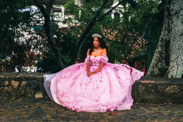 A young woman in a flowing pink dress sits gracefully on a stone wall, surrounded by lush greenery in Colina de San Antonio, Cali.