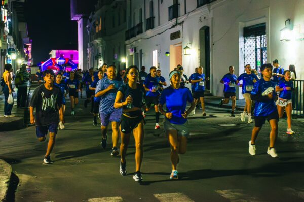 Runners in blue jerseys participate in a nighttime race along the streets of Popayán, Cauca, Colombia, illuminated by city lights.