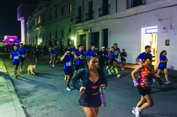 Runners in blue shirts participate in a night race on the streets of Popayán, Cauca, Colombia, with lively urban lights.