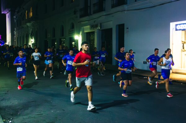 Runners in blue shirts race through the vibrant streets of Popayán, Cauca, Colombia at night, creating an energetic atmosphere.
