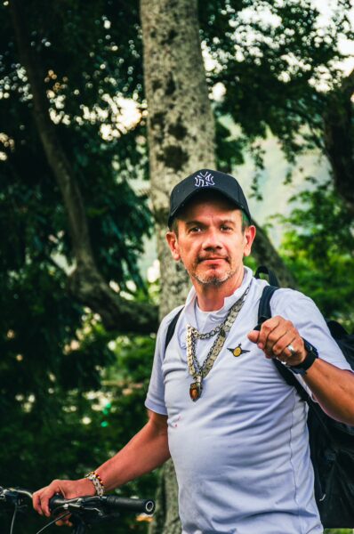 A man wearing a baseball cap and chain stands with a bicycle in lush greenery at Colina de San Antonio, Cali, Valle del Cauca, Colombia.
