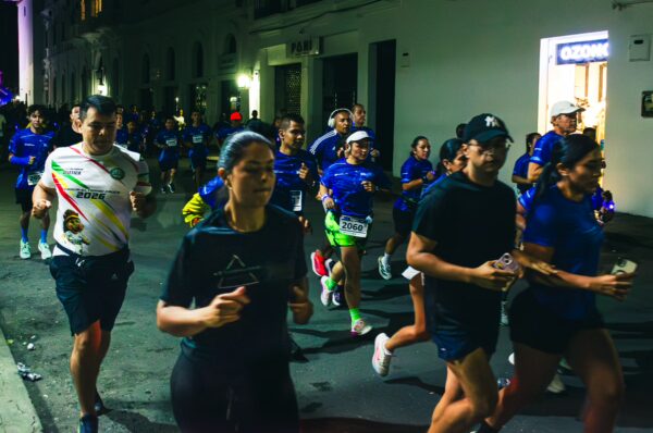 Runners in blue jerseys participate in a lively race through the streets of Popayán, Cauca, Colombia, under vibrant street lights.