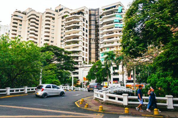 Modern buildings and lush greenery surround a curved road in Cali, Valle del Cauca, Colombia, with people walking nearby.