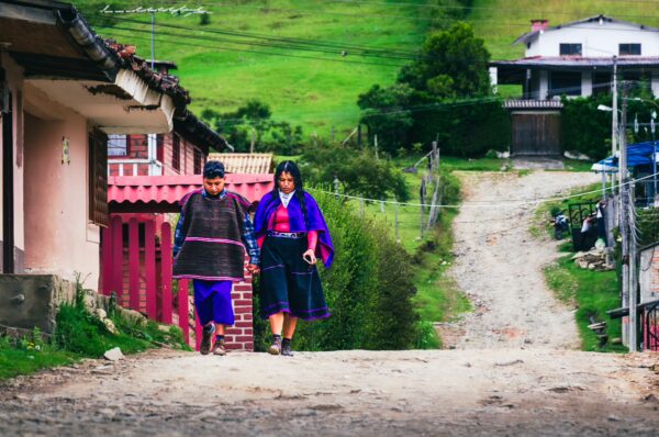 Two people in traditional attire walk down a dirt road lined with greenery, typical of Cauca, Colombia.