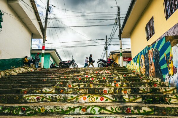 Colorful floral steps lead to a bustling street with motorcycles, against a backdrop of cloudy skies in Cauca, Colombia.