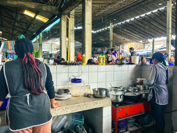 Local women prepare traditional meals in bustling Silva market, colored stalls and goods visible in the background, Cauca, Colombia.