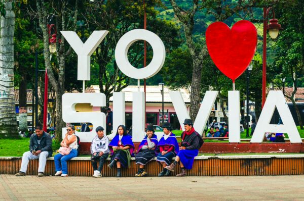 Colorful "Yo <3 Silvia" sign with visitors seated on a bench in Parque Principal Silva, Cauca, Colombia. Lush greenery surrounds the area.
