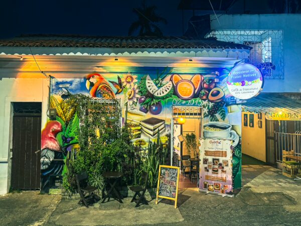 Vibrant street art decorates a café in San Antonio, Cali, with colorful plants and a lively atmosphere at night.