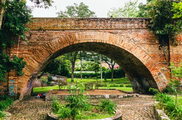 Rustic red brick arch of Puente del Humilladero surrounded by lush greenery and cobblestone pathways in Popayán, Cauca, Colombia.