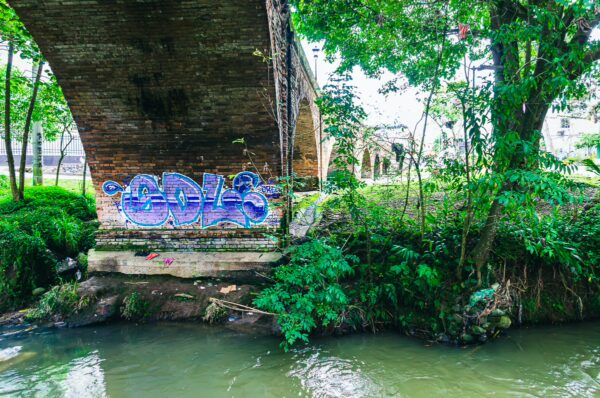 Graffiti adorns the stone base of Puente del Humilladero, surrounded by lush greenery and tranquil water in Popayán, Cauca, Colombia.
