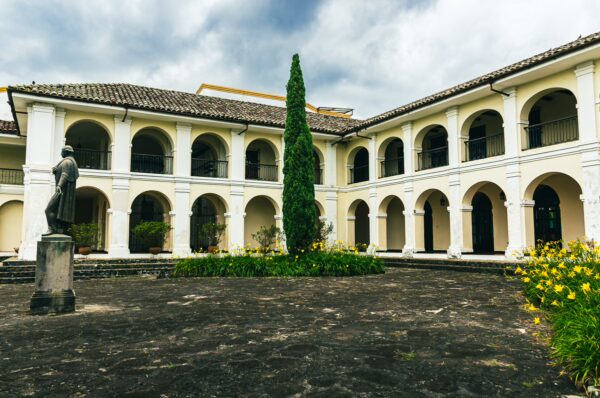 Elegant white arches frame a green courtyard with a tall tree and vibrant yellow flowers in Popayán, Cauca, Colombia.