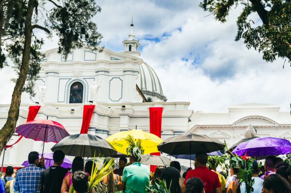 Colorful umbrellas shade a lively crowd in Parque Caldas, with a grand white building and cloudy sky in Popayán, Cauca, Colombia.