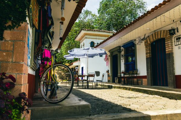 Colorful buildings line the cobblestone street in Pueblito Patojo, featuring a bike and outdoor seating under sun umbrellas in Popayán, Cauca.