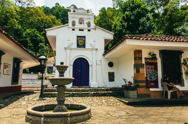 Charming white architecture with a purple door and a central fountain, surrounded by lush greenery in Pueblito Patojo, Popayán.