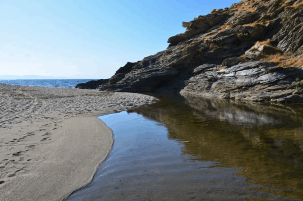Serene Kallianos Beach features golden sand, clear water, and rugged rock formations under a bright blue sky.