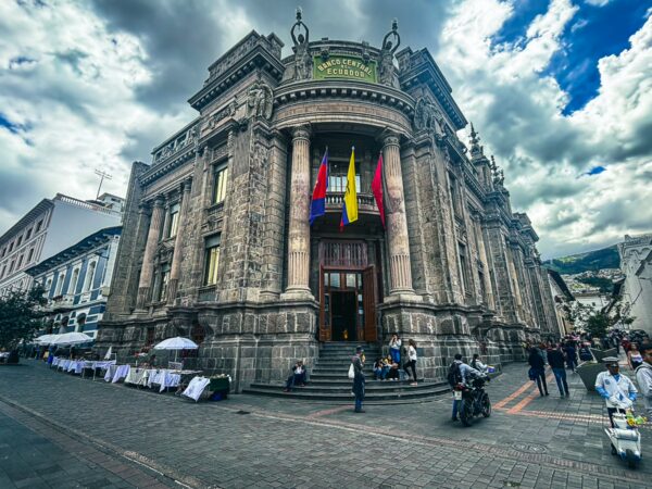 Banco Central in Quito, Ecuador features ornate architecture with colorful flags and bustling street vendors under a dramatic sky.