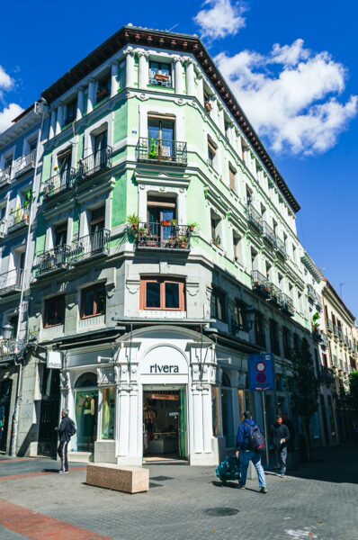 Charming green building with balconies and shopfront on a sunny street in Madrid, Spain, surrounded by clear blue skies.