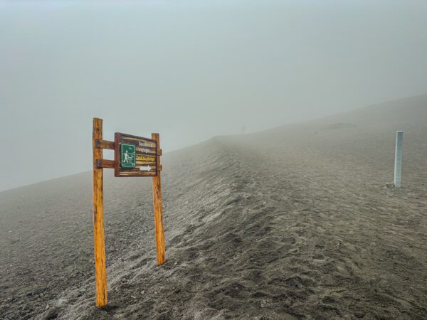 Fog envelops a sandy trail leading up the slopes of Mount Cotopaxi in Ecuador, marked by a weathered wooden sign.