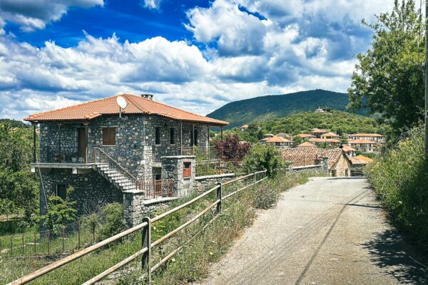 Charming stone houses line the road in Nymfasia, Arcadia, Greece, with vibrant greenery and a backdrop of rolling hills.
