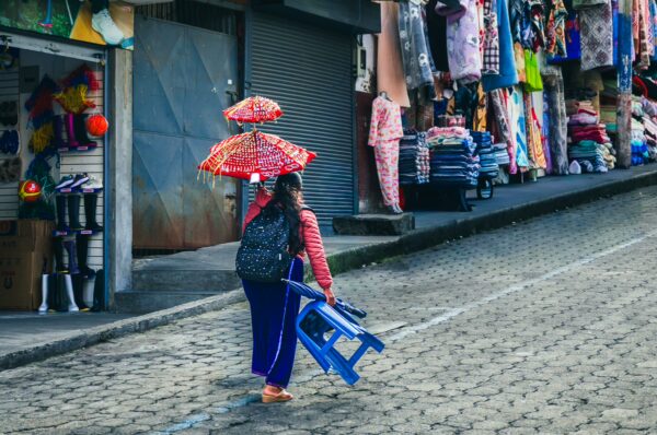 Vibrant colors fill the market in Tulcán, Ecuador, as a girl carries a blue chair while shielding herself with red umbrellas.