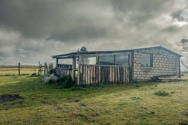 Rustic building with a wooden fence and overcast sky in Cotopaxi National Park, Ecuador, surrounded by lush grasslands.