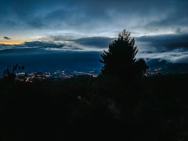 Twilight view from Imbabura Volcano, Ecuador, with distant lights and silhouette of a coniferous tree against a dramatic sky.
