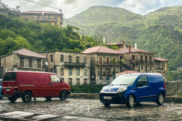 Colorful vans parked in the rain near stone buildings, surrounded by lush green mountains in Lagadia, Arcadia, Greece.