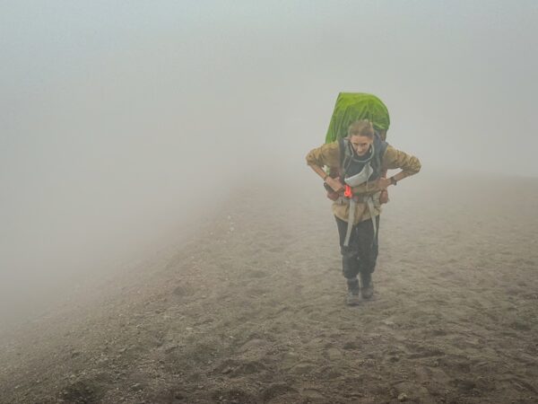 Hiker navigating through thick fog with a green backpack on the sloping terrain of Mount Cotopaxi, Ecuador.