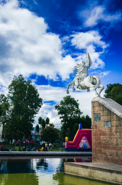 Elegant white statue of a horseman overlooks the serene water of Parque Ayora, with vibrant greens and playful structures in Tulcán, Ecuador.