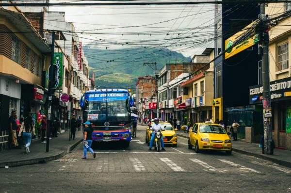 Busy street scene in Ibarra, Ecuador, featuring a blue bus, yellow taxis, and shops, with green mountains in the background.