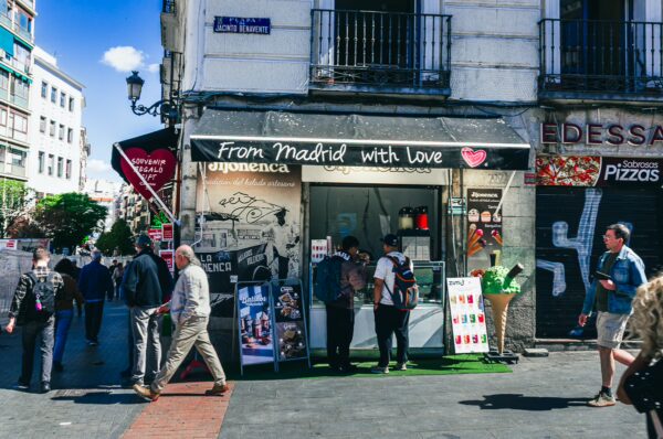 Colorful storefront labeled "From Madrid with Love" in a bustling Madrid street, surrounded by pedestrians and vibrant city life.