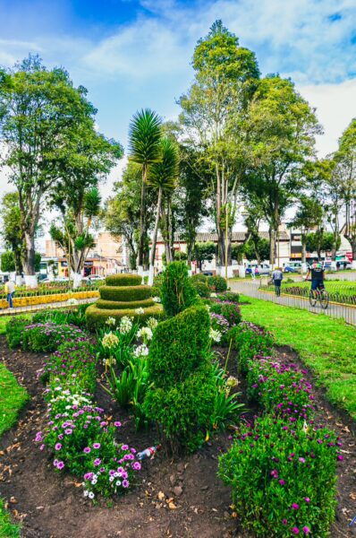 Lush greenery and vibrant flower beds create a picturesque scene in Parque Ayora, Tulcán, Ecuador, under a bright blue sky.