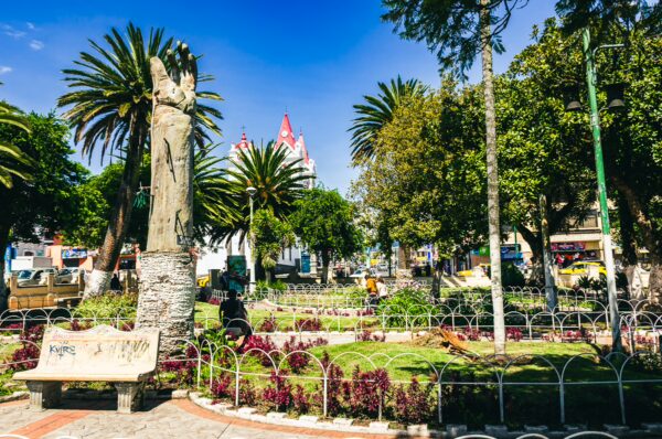 Lush greenery and vibrant flowers surround a central statue in Central Park, Machachi, Ecuador, under a clear blue sky.