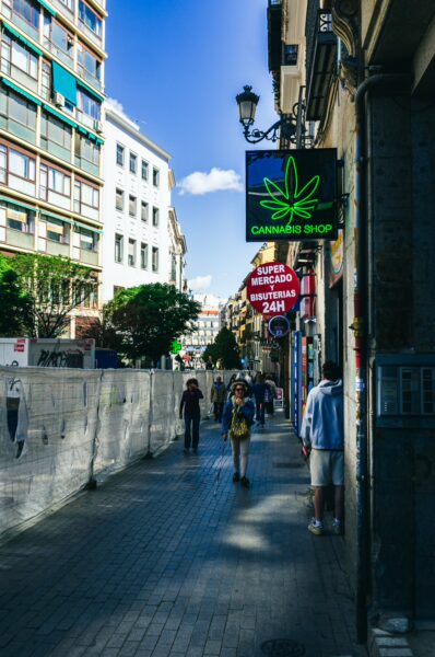 Lively street scene in Madrid featuring shops, pedestrians, and a distinctive cannabis shop sign under a clear blue sky.