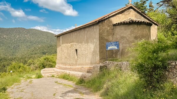 Stone chapel nestled in lush greenery under a bright blue sky, set against the backdrop of the Arcadian mountains in Magouliana, Greece.