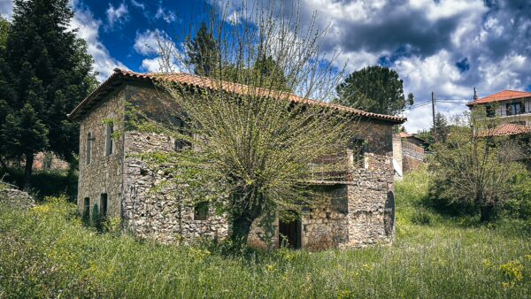 Rustic stone house surrounded by vibrant greenery and flowering plants in Nymfasia, Arcadia, Greece, under a cloudy sky.