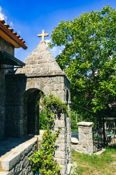 Panagia Kernitsis Monastery features a stone archway, a cross atop the tower, and lush greenery under a bright blue sky in Arcadia, Greece.