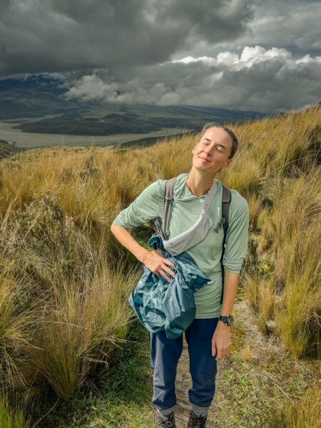 Hiker stands amidst tall grass on a trail, with dark clouds gathering over the volcanic landscape of Ruminahui, Ecuador.