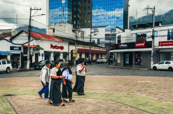 Traditional dressed locals walk through a bustling plaza in Ibarra, Ecuador, with modern buildings in the background.