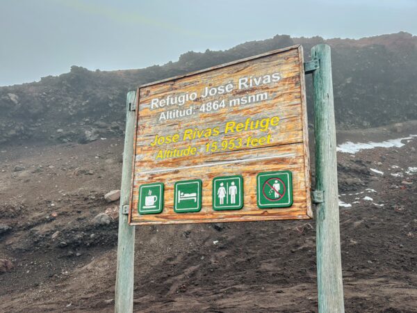Wooden sign for Jose Rivas Refuge at Mount Cotopaxi, Ecuador, displaying altitude and icons for facilities amidst rocky terrain.