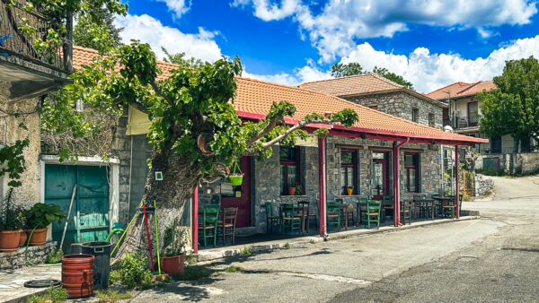 Charming stone buildings line the street in Nymfasia, Arcadia, Greece, with a vibrant café shaded by a leafy vine.