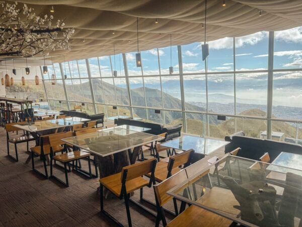 Stylish café with large windows offering panoramic views of Pichincha Volcano, featuring wooden tables and a modern design.