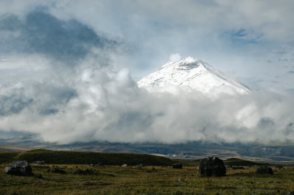 Majestic snow-capped Cotopaxi peak rises above lush green fields, framed by clouds in Cotopaxi National Park, Ecuador.